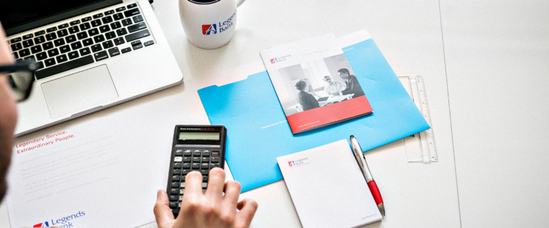 An photo of an above view of a workspace with a person using a calculator next to a laptop, cup of coffee, and Legend's Bank folder / papers.