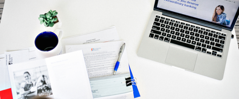 A photo of an above view of a workspace with Legend's Bank papers and checks next to a laptop with the Legends Bank website on screen.